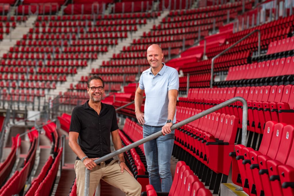 Twee mannen staan in een stadion tussen rode tribuneplaatsen.