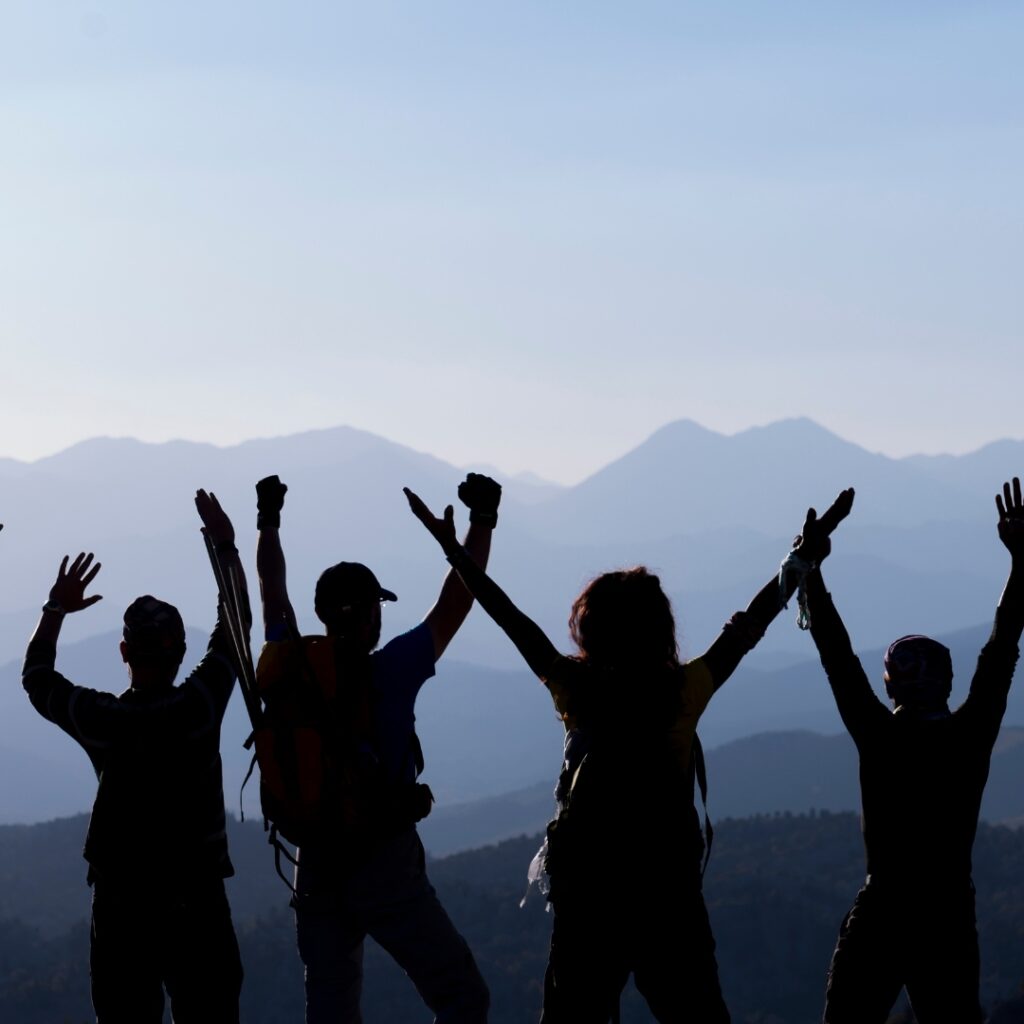 A group of hikers raises their arms in celebration against a mountainous backdrop.