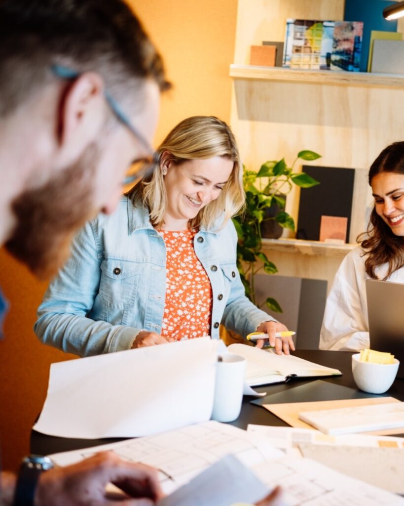 Drie collega’s bespreken documenten en werken samen aan laptop in moderne kantooromgeving.