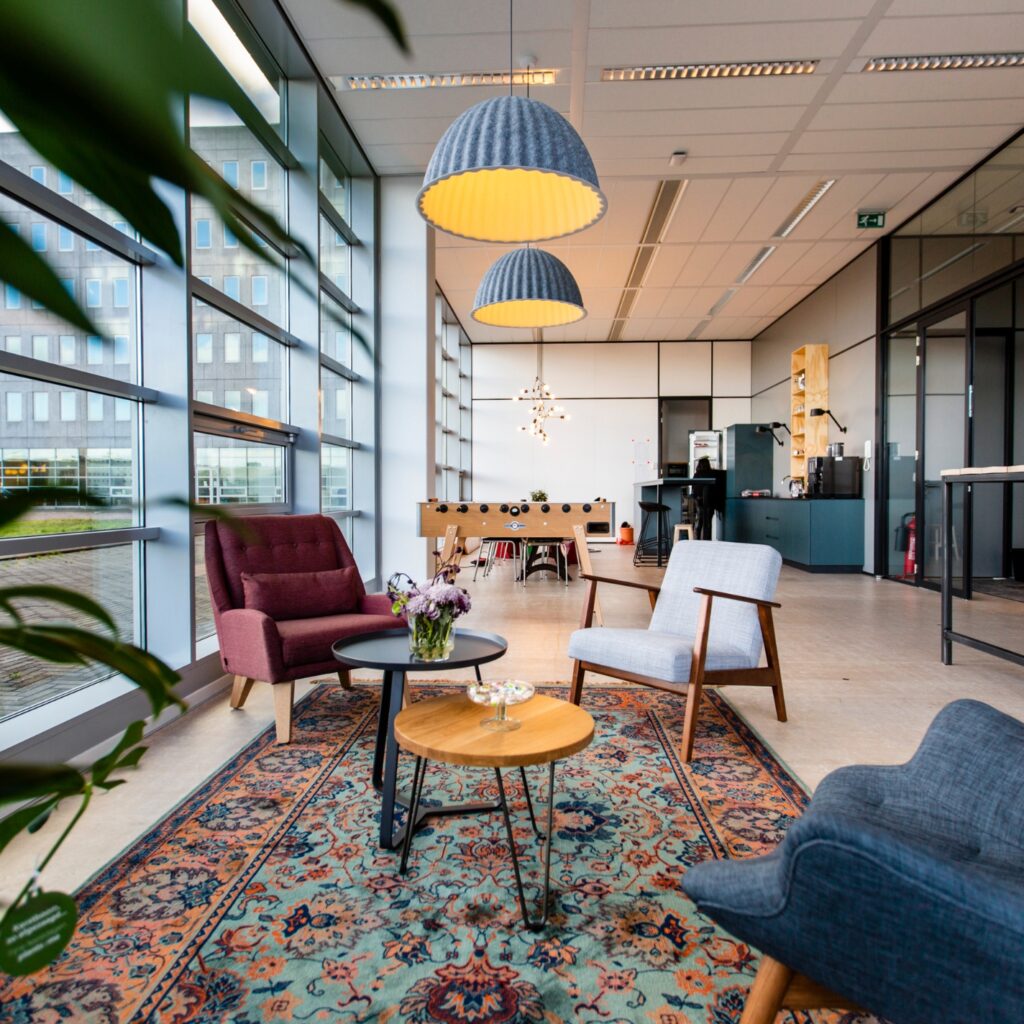 Informele kantoorruimte met comfortabele stoelen, vloerkleed en grote ramen in een lichte werkomgeving.