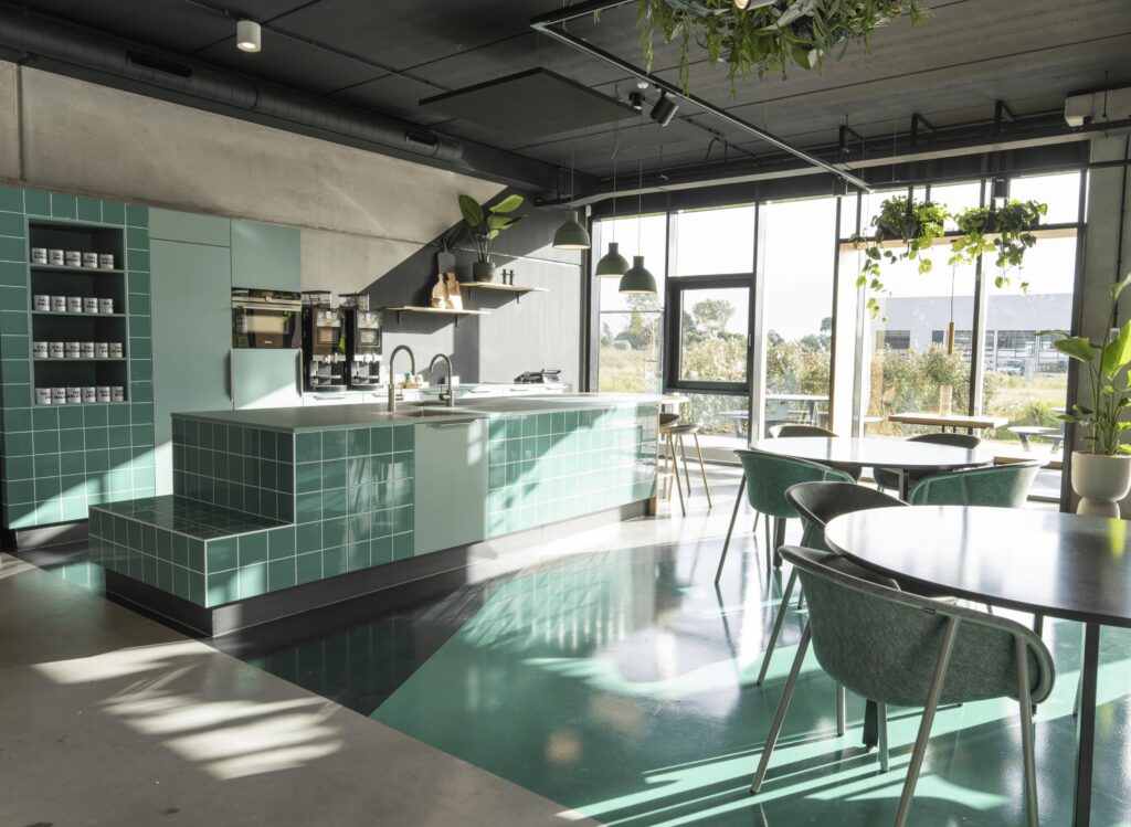 Een moderne kantoorpantry met een groen betegeld kookeiland en ingebouwde apparatuur. Rondom staan ronde tafels met groene stoelen bij grote ramen die veel daglicht binnenlaten. Het plafond heeft een industriële afwerking met zichtbare leidingen en hangpl