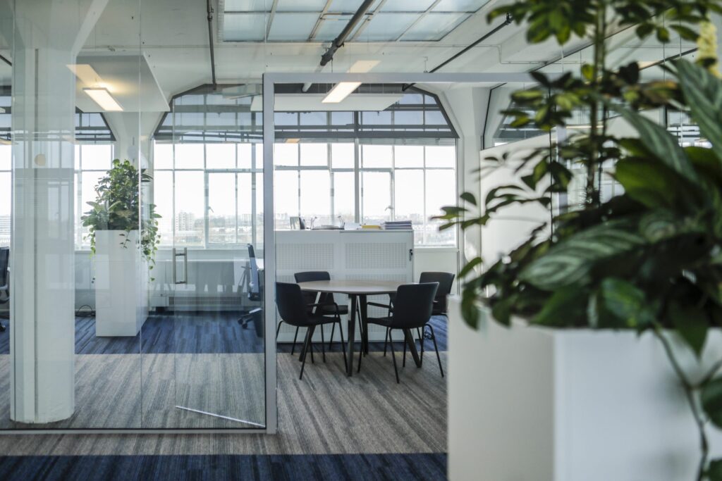 Een moderne kantoorruimte met een glazen vergaderruimte waarin een ronde tafel en vier stoelen staan. Grote ramen zorgen voor veel daglicht en uitzicht naar buiten. Op de voorgrond is een plant in een witte pot zichtbaar en het plafond heeft een industrië