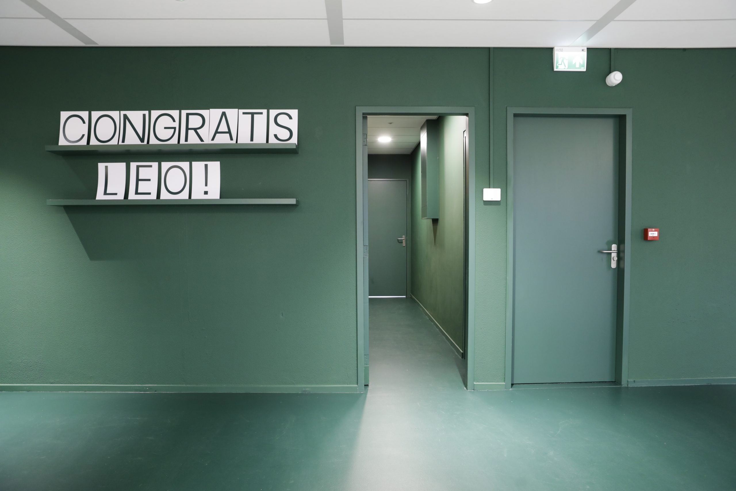 Een groene gang in een kantoorruimte met twee deuren aan de rechterzijde. Aan de linkerwand hangt een plank met blokletters waarop “CONGRATS LEO!” staat. De vloer en muren zijn in dezelfde groene tint afgewerkt.