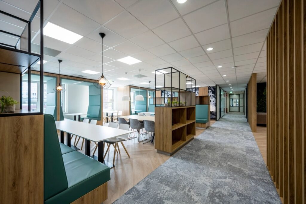 Een moderne kantoorruimte met verschillende zit- en werkplekken langs een lange gang. Links staan tafels met stoelen en groene zitbanken, gescheiden door houten kasten en glazen wanden. Rechts loopt een brede gang met grijze vloerbedekking en meerdere deu