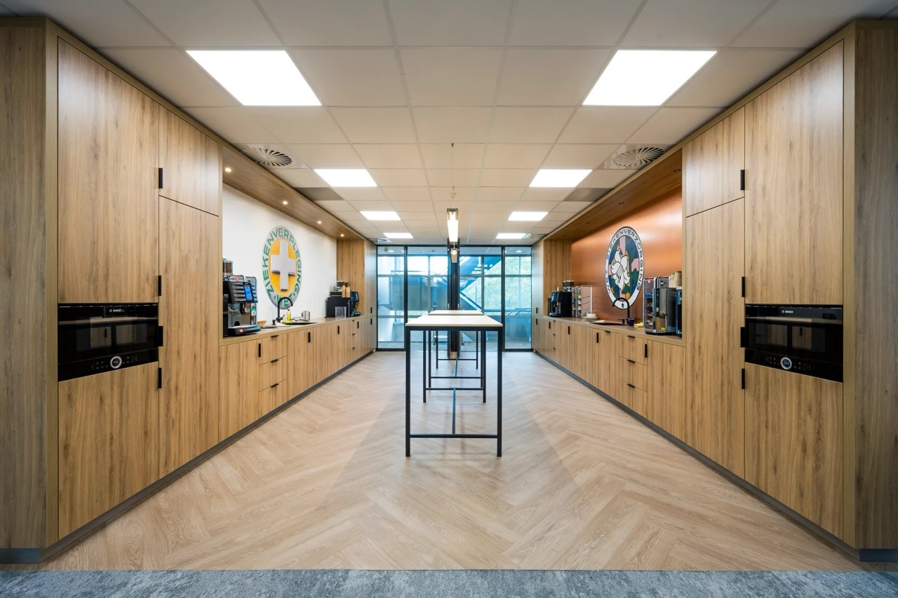 Een moderne kantoorpantry met houten kastenwanden aan beide zijden en ingebouwde koffieapparaten. In het midden staat een hoge tafel met krukken. Aan het einde van de ruimte zorgen grote ramen voor veel natuurlijk licht.