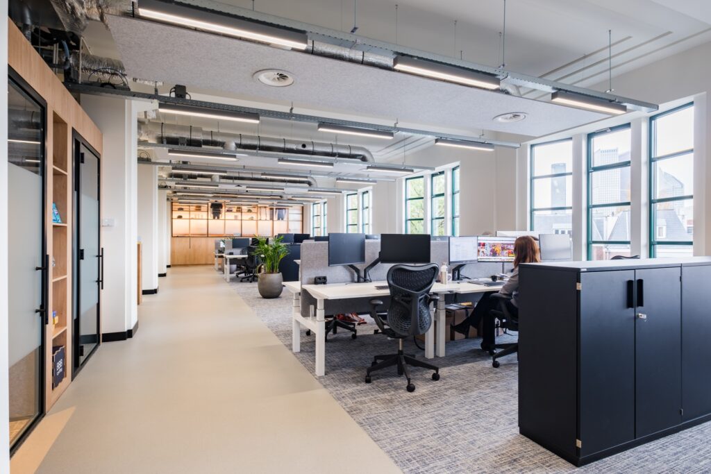 Open kantoorruimte met bureaus, ergonomische stoelen en grote ramen die zorgen voor veel daglicht, met medewerkers aan het werk.