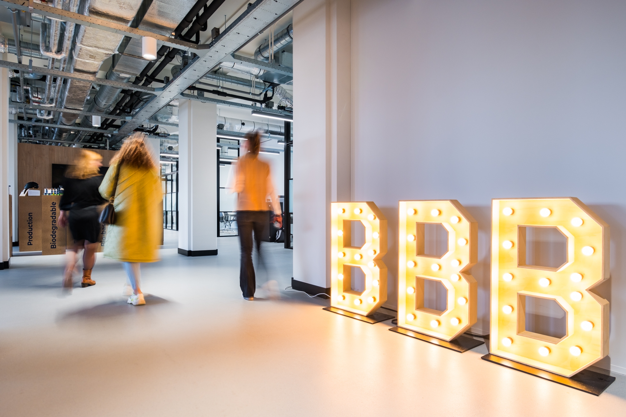 In een modern kantoor met industriële plafonds lopen drie personen door een open ruimte. Aan de rechterkant staan grote verlichte letters “BBB” tegen de muur, die warm licht uitstralen.