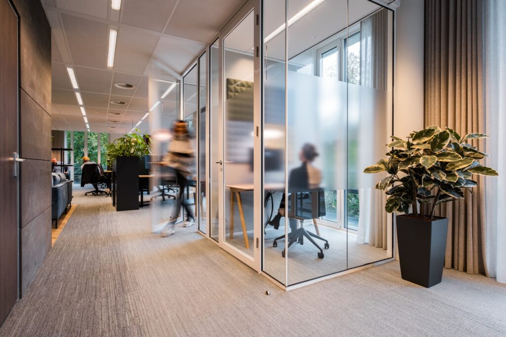 Moderne kantoorruimte met glazen vergaderruimtes, waarbij medewerkers werken en een persoon langsloopt in de gang.