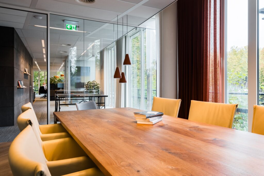 Een moderne vergaderruimte met een grote houten tafel en gele stoelen. Op de tafel liggen enkele boeken. Naast de tafel hangen drie zwarte hanglampen. Achter de glazen wand is een andere vergaderruimte zichtbaar en grote ramen zorgen voor veel natuurlijk