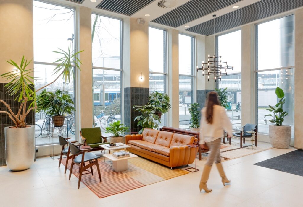 Moderne kantoorruimte met zitnissen en gele frameconstructies waar collega’s werken en overleggen. Twee mensen voeren een gesprek aan een tafel met laptop, terwijl op de achtergrond iemand rustig een krant leest, wat de informele en flexibele werkomgeving