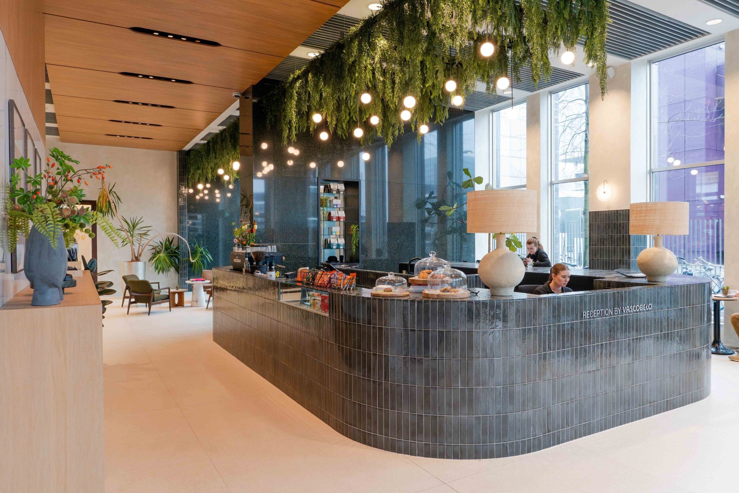 Een moderne vergaderruimte met een ronde houten tafel en bruine stoelen. Aan de muur hangt een groot kunstwerk en via de hoge ramen is uitzicht op kantoorgebouwen en groen in de stad. De ruimte is licht en strak ingericht met een rustige uitstraling.