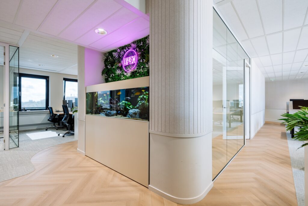 Een moderne kantoorruimte met een ingebouwd aquarium in een lichte wand. Boven het aquarium hangt een paarse neonverlichting tegen een groene plantenwand. Rechts is een glazen vergaderruimte zichtbaar en de vloer heeft een licht houten visgraatpatroon.