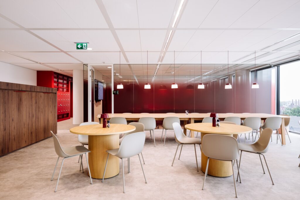Een moderne kantineruimte met ronde houten tafels en lichtgekleurde stoelen. Aan de achterzijde bevindt zich een glazen wand met een rode achtergrond en hanglampen erboven. Links zijn rode lockers zichtbaar en het plafond is voorzien van inbouwverlichting