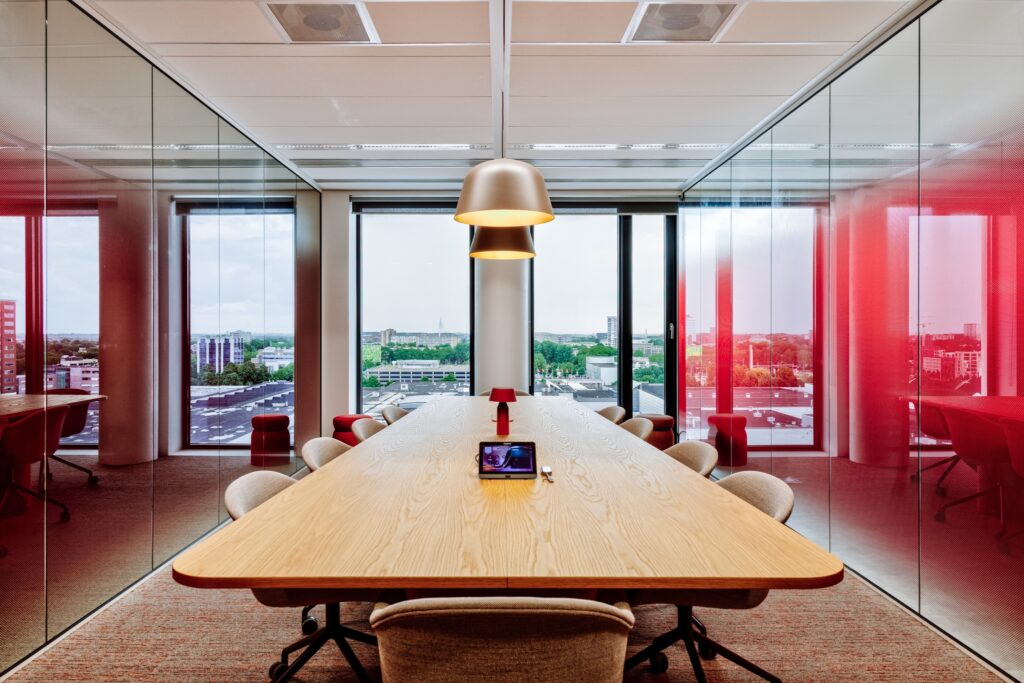 Moderne vergaderruimte met een lange houten tafel en beige stoelen, omringd door glazen wanden met rode accenten, met uitzicht op een stedelijk landschap door grote ramen.