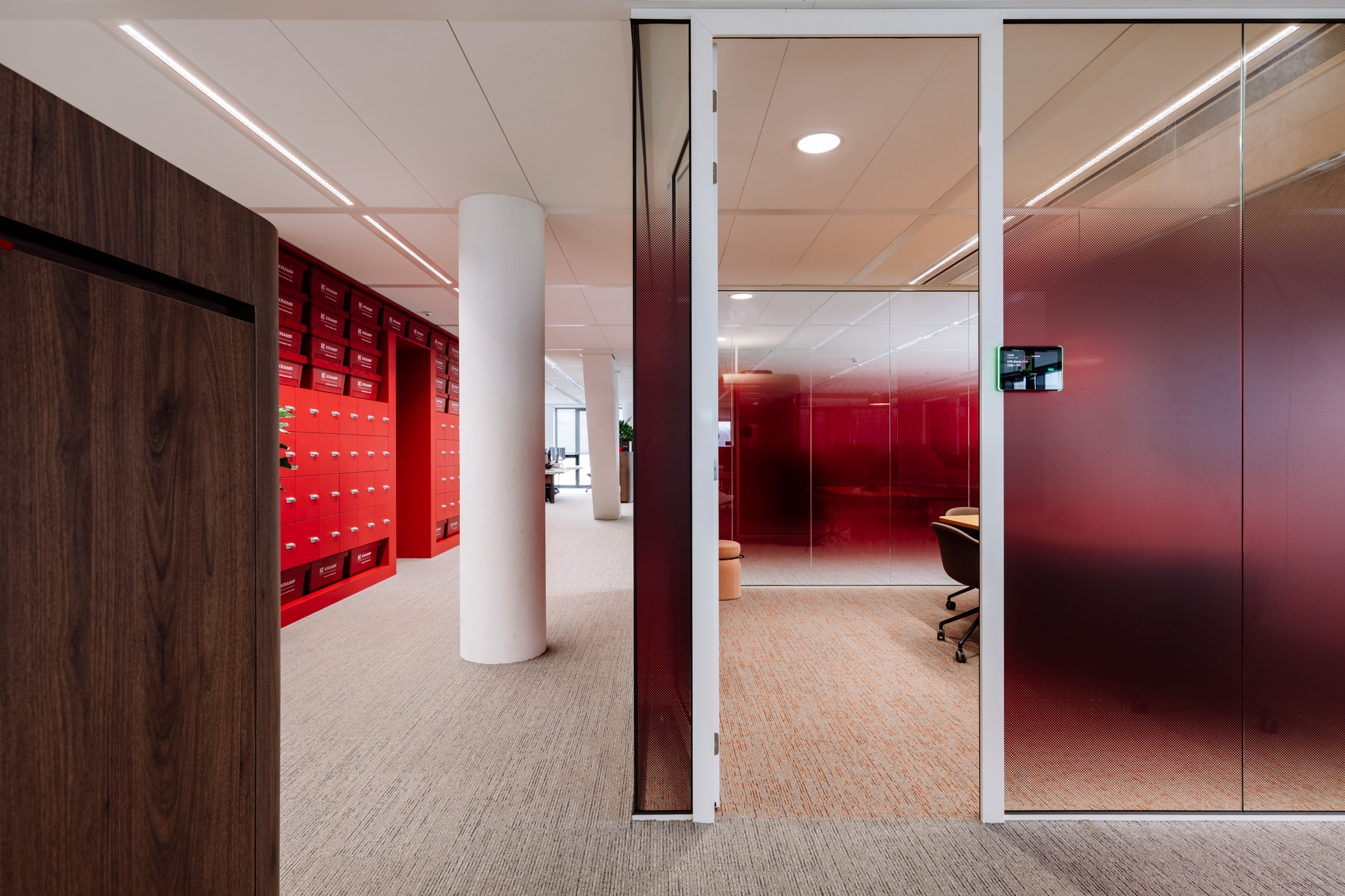 Een moderne kantoorruimte met een gang waarin rode lockers langs de muur staan. Rechts bevindt zich een glazen wand met een rood getinte afwerking en daarachter een vergaderruimte met bureaustoelen. De ruimte heeft een lichte vloer en een strak afgewerkt