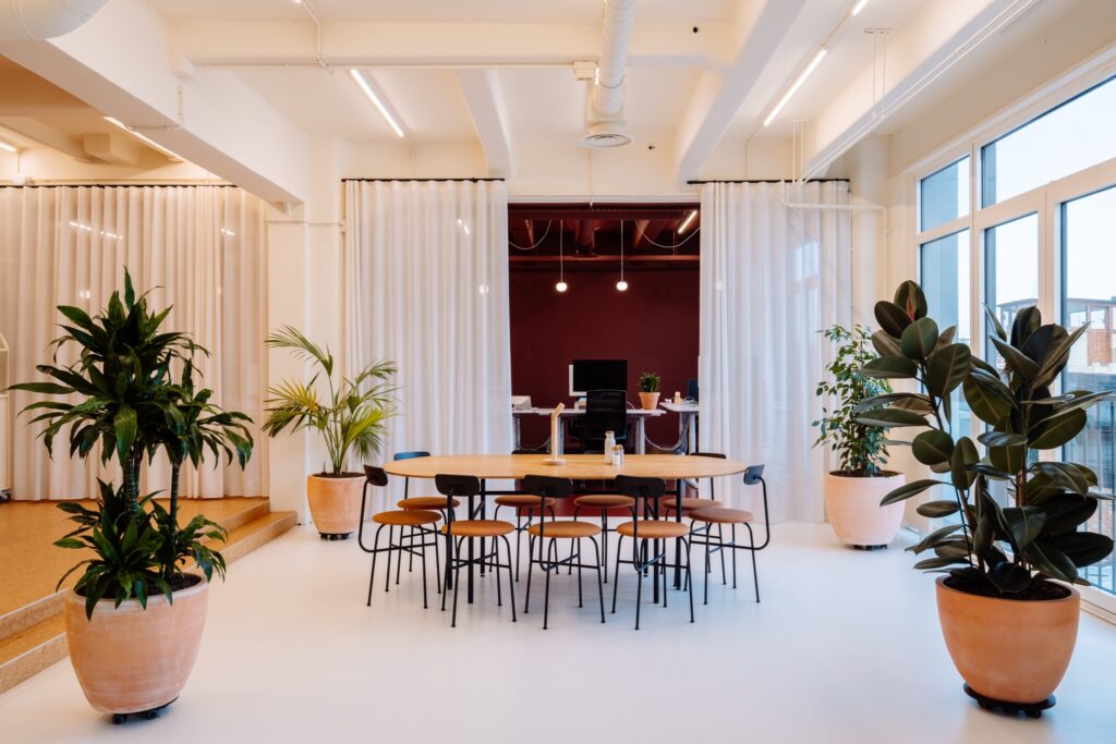 Moderne kantoorruimte met ronde vergadertafel, stoelen, planten en lichte inrichting met gordijnen en werkplekken op de achtergrond.
