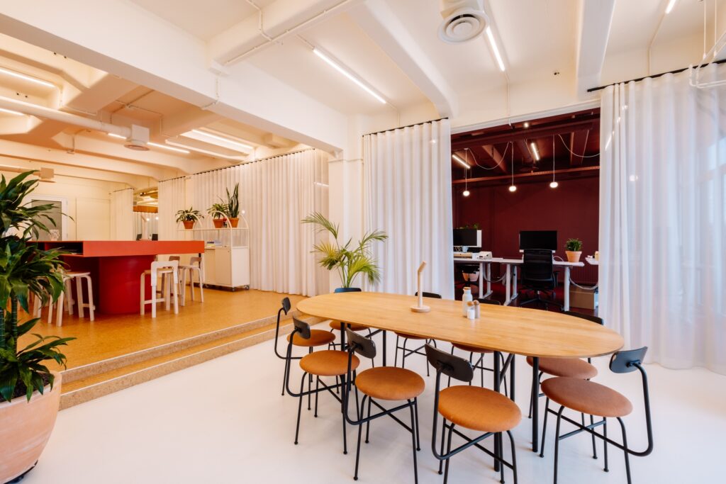 Moderne kantoorruimte met een lange houten tafel en krukken op de voorgrond, een verhoogde bar met witte barkrukken en een werkruimte me