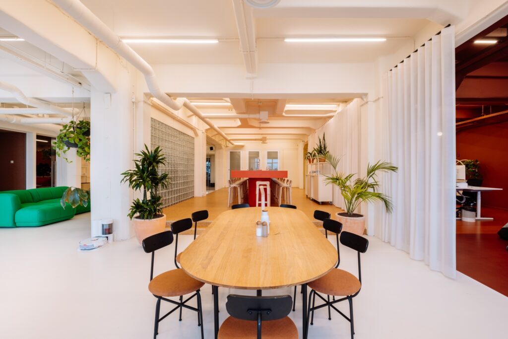 Moderne, lichte vergaderruimte met een lange houten tafel, zwarte stoelen, groene planten en witte gordijnen in een open kantooromgeving.