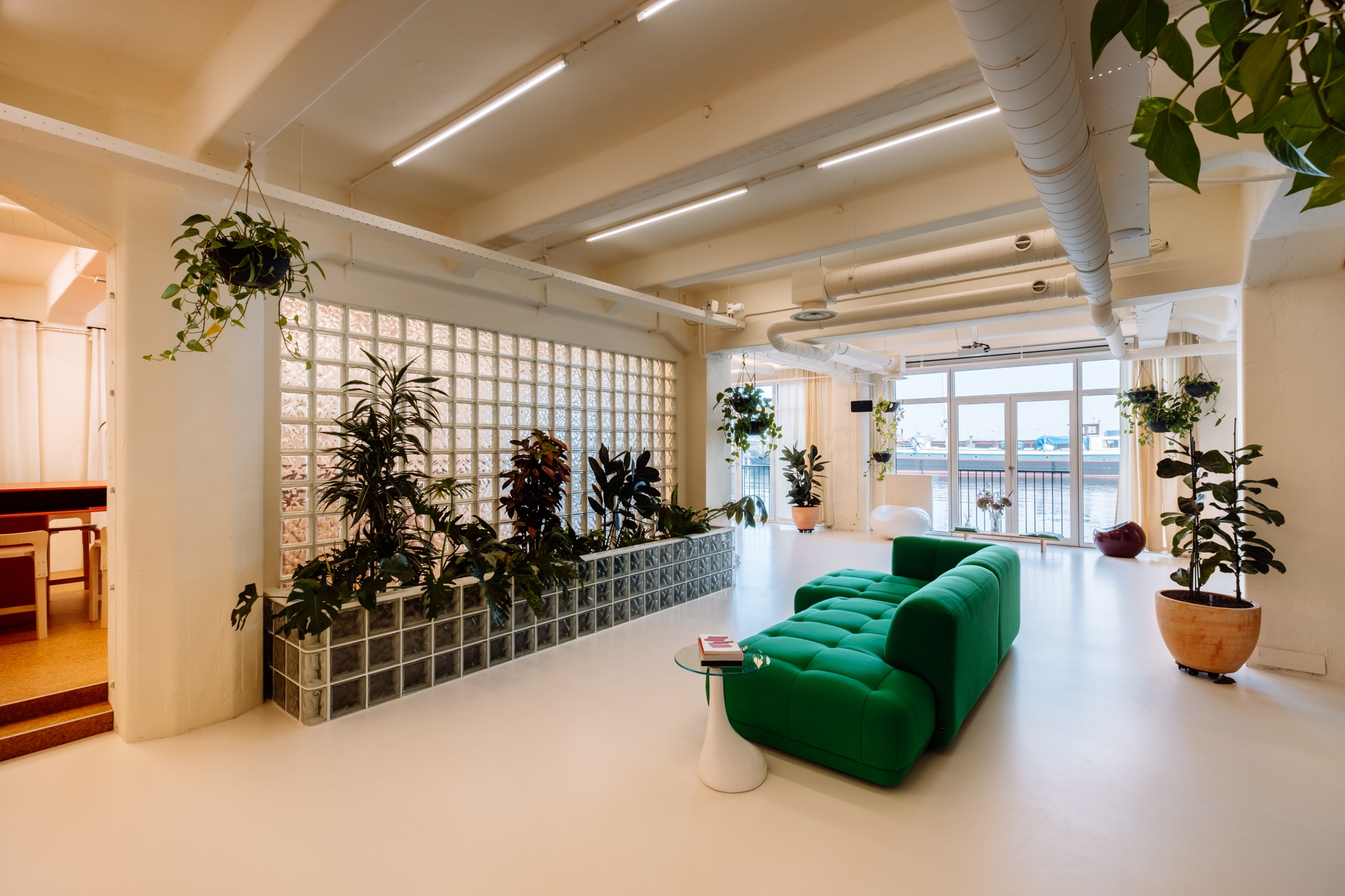 Een ruime, lichte kantoorruimte met witte vloeren en plafonds, voorzien van grote ramen met uitzicht naar buiten. In het midden staat een groene bank met een klein rond tafeltje ervoor. Langs een halfhoge wand van glasblokken staan meerdere grote planten,