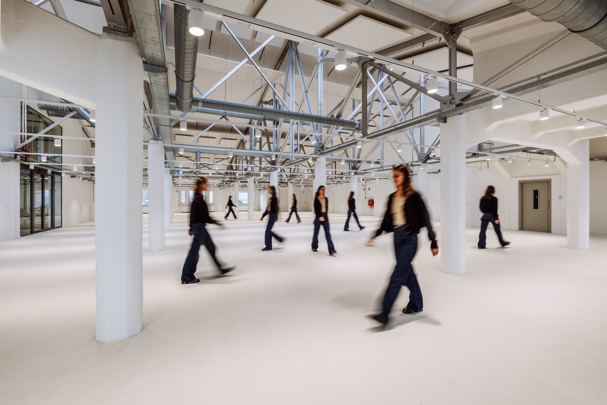 Een grote, open industriële ruimte met witte kolommen en zichtbare stalen constructies en ventilatiebuizen aan het plafond. Meerdere personen lopen door de ruimte, deels onscherp door beweging. De vloer is licht en de ruimte oogt ruim en minimalistisch.