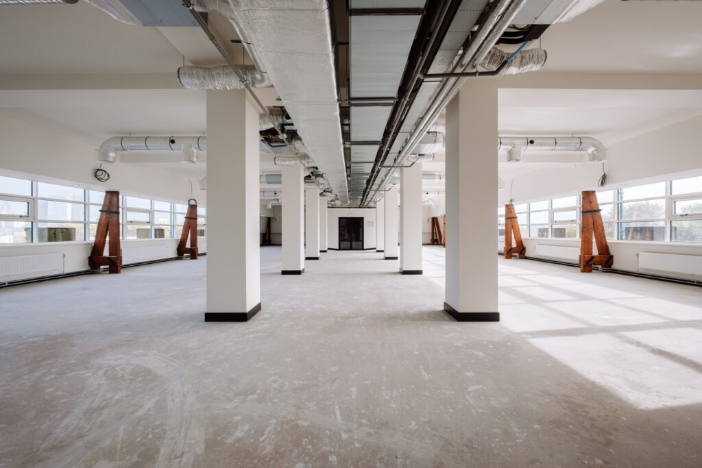 Een grote, lege kantoorruimte met betonnen vloer en witte kolommen in een rij. Aan het plafond zijn ventilatiekanalen en leidingen zichtbaar. Grote ramen aan beide zijden zorgen voor veel daglicht in de open ruimte.