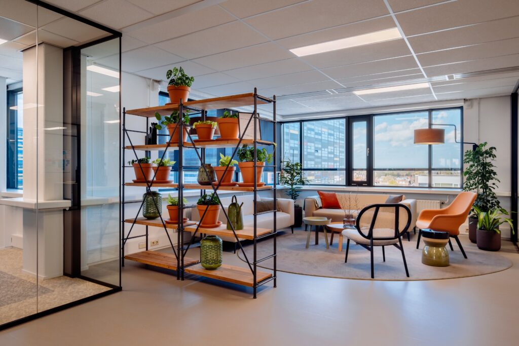 Moderne kantoorruimte met open kast vol planten, zithoek met banken en stoelen, en grote ramen met uitzicht op de stad.