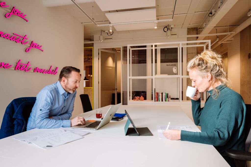 Twee collega’s werken samen aan een bureau, met laptops en documenten in een moderne kantoorruimte met glazen wanden.