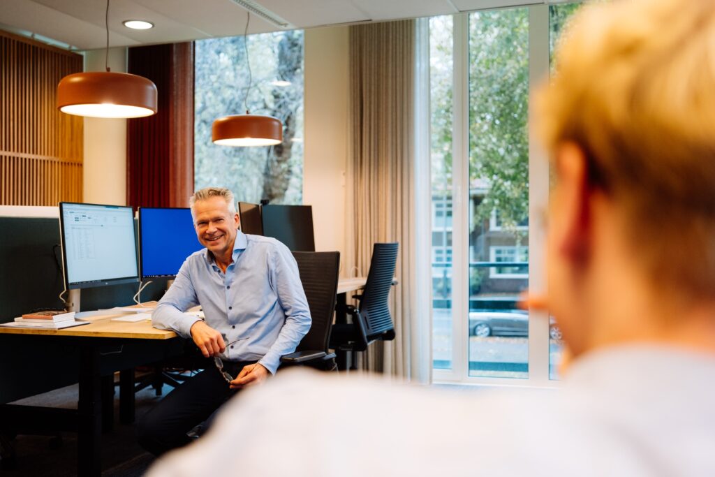 Man in gesprek in moderne kantoorruimte met grote ramen en werkplekken.