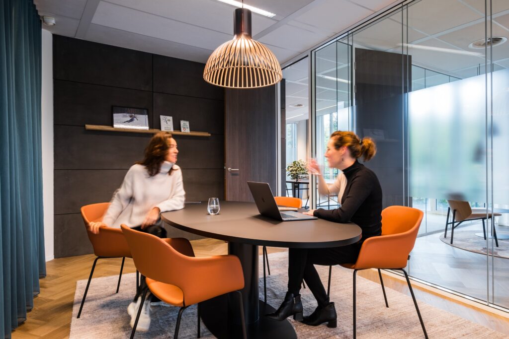 Twee vrouwen in overleg aan ronde tafel in moderne vergaderruimte met glazen wanden.