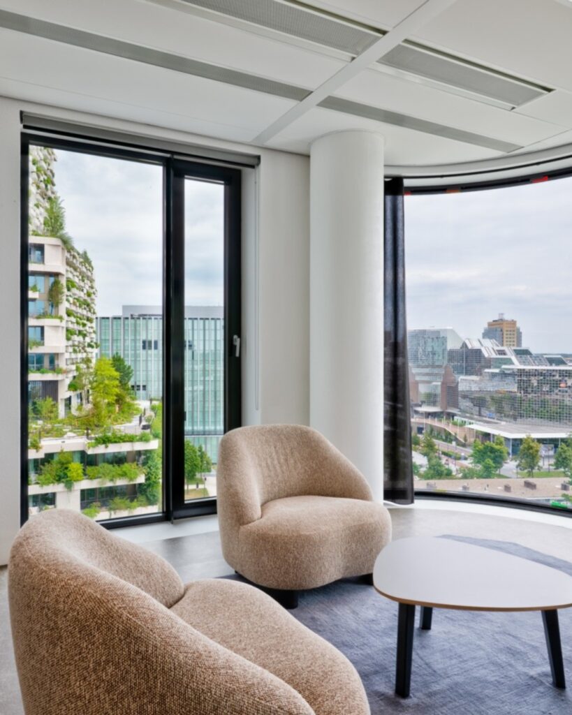 Moderne zithoek met comfortabele stoelen, grote ramen en uitzicht op een groene stedelijke omgeving met moderne gebouwen.