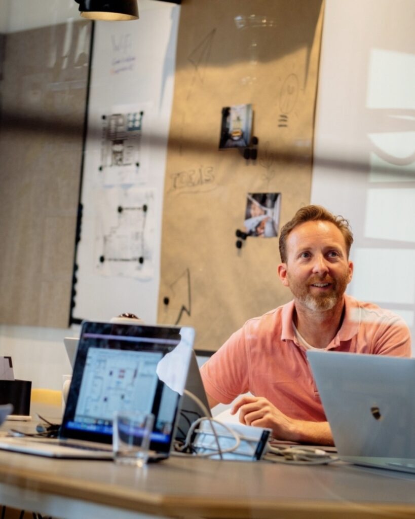 Medewerker zit aan een vergadertafel met laptops en kijkt aandachtig op tijdens een overleg. Op de achtergrond hangen schetsen en notities op een glazen wand, wat een creatieve en samenwerkende werkomgeving laat zien.