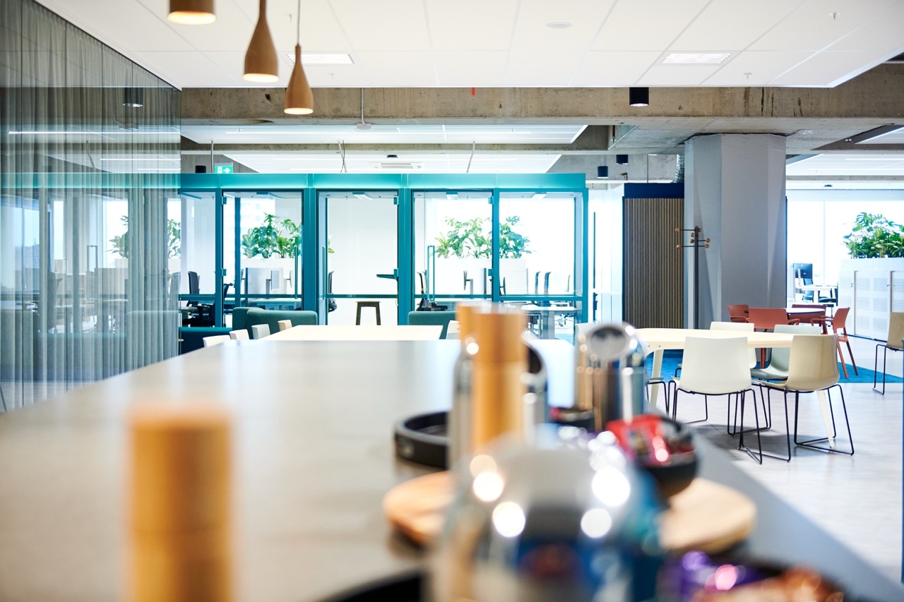 Een moderne kantoorruimte met een lange tafel op de voorgrond waarop dienbladen en bekers staan. Op de achtergrond zijn glazen deuren met turquoise kozijnen zichtbaar en verschillende tafels en stoelen in een open ontmoetingsruimte. De ruimte is licht en