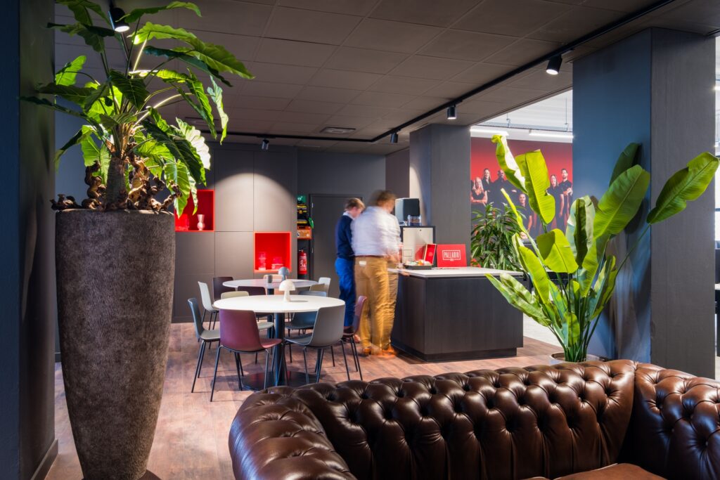 Een moderne kantoorruimte met een zithoek en ronde tafel met stoelen. Op de achtergrond staan twee personen bij een balie. Grote planten en rode nissen in de muur geven de ruimte een warme uitstraling. Op de voorgrond is een donkerbruine leren bank zichtb
