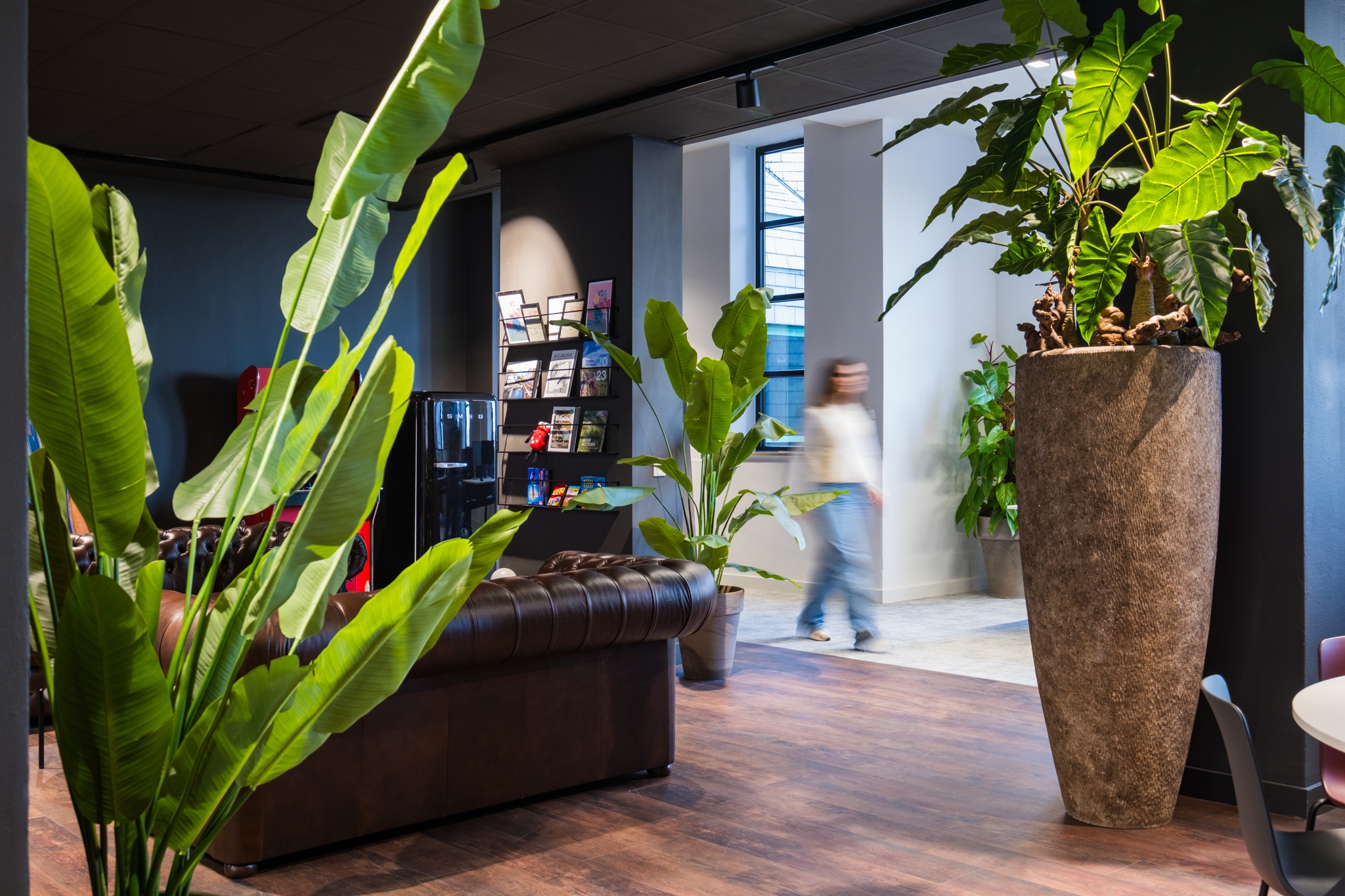 Een moderne kantoorruimte met een donkerbruine leren bank en grote planten in hoge potten. Op de achtergrond staat een boekenkast met tijdschriften en loopt een persoon onscherp door de ruimte. De vloer is van hout en de ruimte heeft een warme, eigentijds