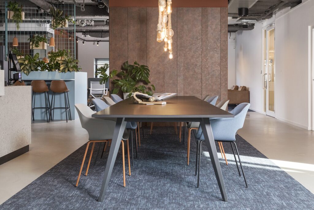 Een moderne vergaderruimte met een grote donkere tafel en grijze stoelen eromheen. Boven de tafel hangt een designlamp met zichtbare gloeilampen. Op de achtergrond zijn planten en een industriële kantooromgeving zichtbaar met een open plafond en metalen d
