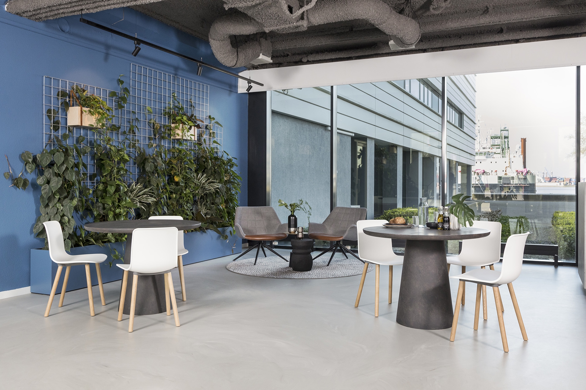Een moderne kantoorruimte met ronde tafels en witte stoelen bij grote ramen met uitzicht naar buiten. Links staat een blauwe wand met klimplanten en groene beplanting. De ruimte heeft een open, industriële uitstraling met zichtbare ventilatiebuizen aan he
