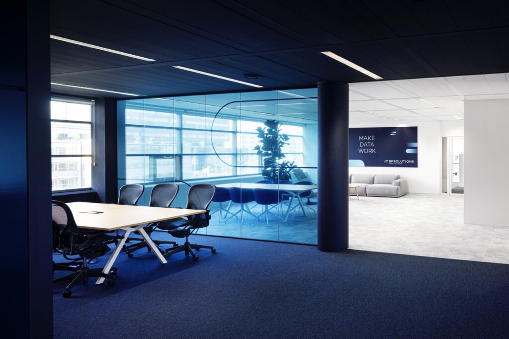 Een moderne kantoorruimte met een vergadertafel en stoelen op een donker tapijt. Rechts bevindt zich een glazen vergaderruimte met blauwe tint. Op de achterwand staat de tekst “MAKE DATA WORK” boven een lichte zithoek. De ruimte heeft een strak plafond me