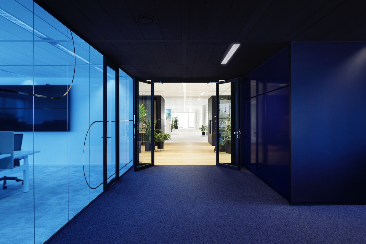 Een moderne kantoorruimte met een donkere gang en glazen vergaderruimtes met blauwe tint aan de linkerzijde. Dubbele glazen deuren leiden naar een lichtere ruimte met planten en zitplekken. Het plafond heeft strakke verlichting en de vloer is bekleed met