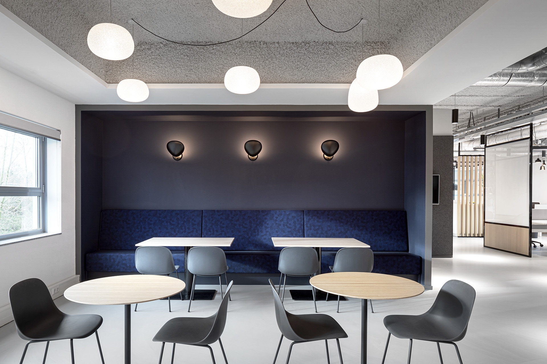 Een moderne zithoek in een kantoor met ronde tafels en grijze stoelen. Tegen de achterwand staat een lange blauwe bank onder drie wandlampen. Aan het plafond hangen meerdere ronde plafondlampen. De ruimte heeft een rustige en minimalistische uitstraling.