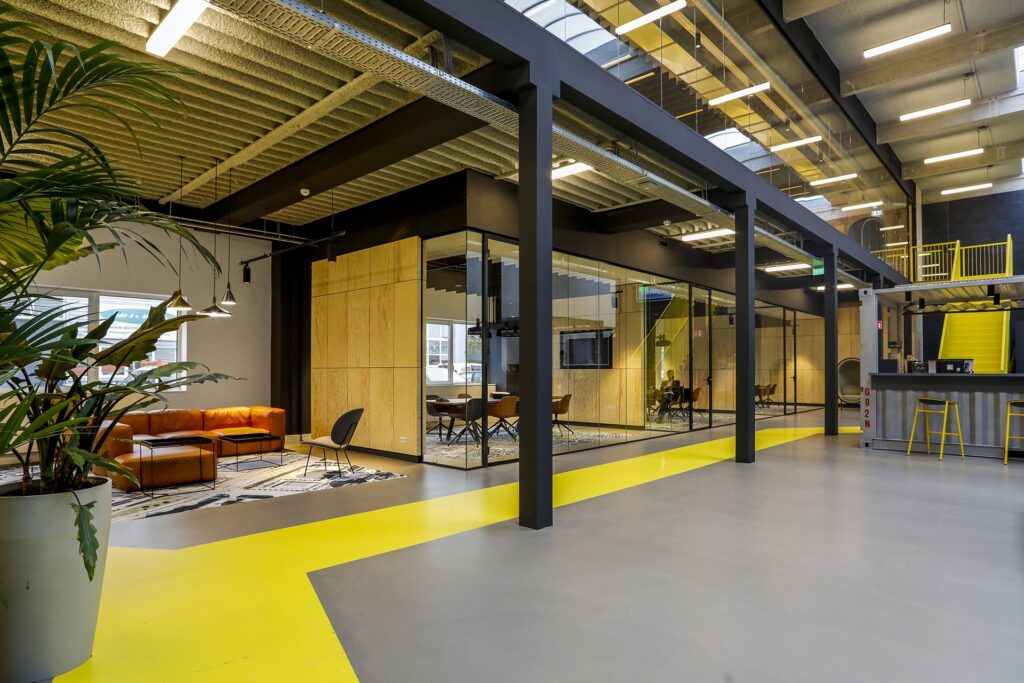 Een moderne kantoorruimte met een industriële uitstraling, zwarte stalen kolommen en een plafond met zichtbare constructie. In het midden bevindt zich een glazen vergaderruimte met houten wanden. Links staat een zithoek met een oranje bank en stoelen. Ove