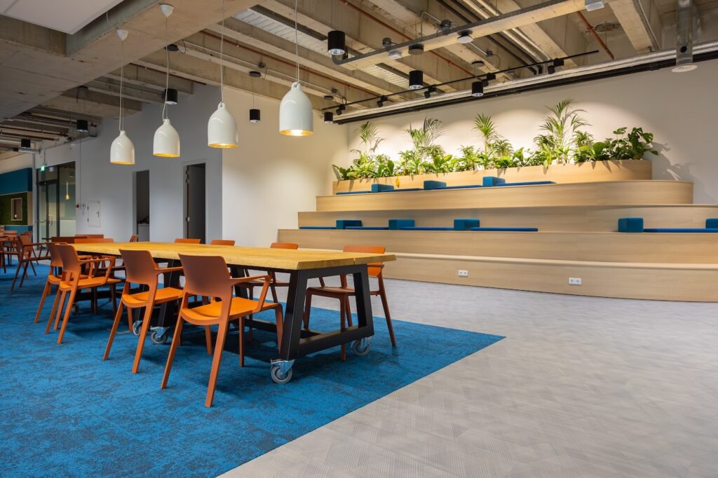 Een moderne kantoorruimte met een grote tafel op wielen en oranje stoelen op een blauw vloerkleed. Aan de achterzijde bevindt zich een houten tribune met geïntegreerde zitplaatsen en planten bovenop. Boven de ruimte hangen witte hanglampen en het plafond