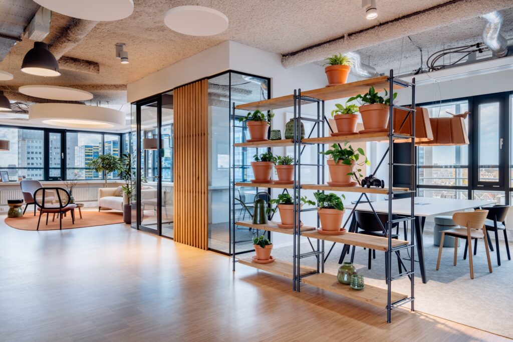 Een moderne kantoorruimte met een open stellingkast gevuld met terracotta potten en groene planten. Op de achtergrond staat een glazen vergaderruimte met zwarte frames en houten wandpanelen. De ruimte heeft een lichte houten vloer en een warm, eigentijds
