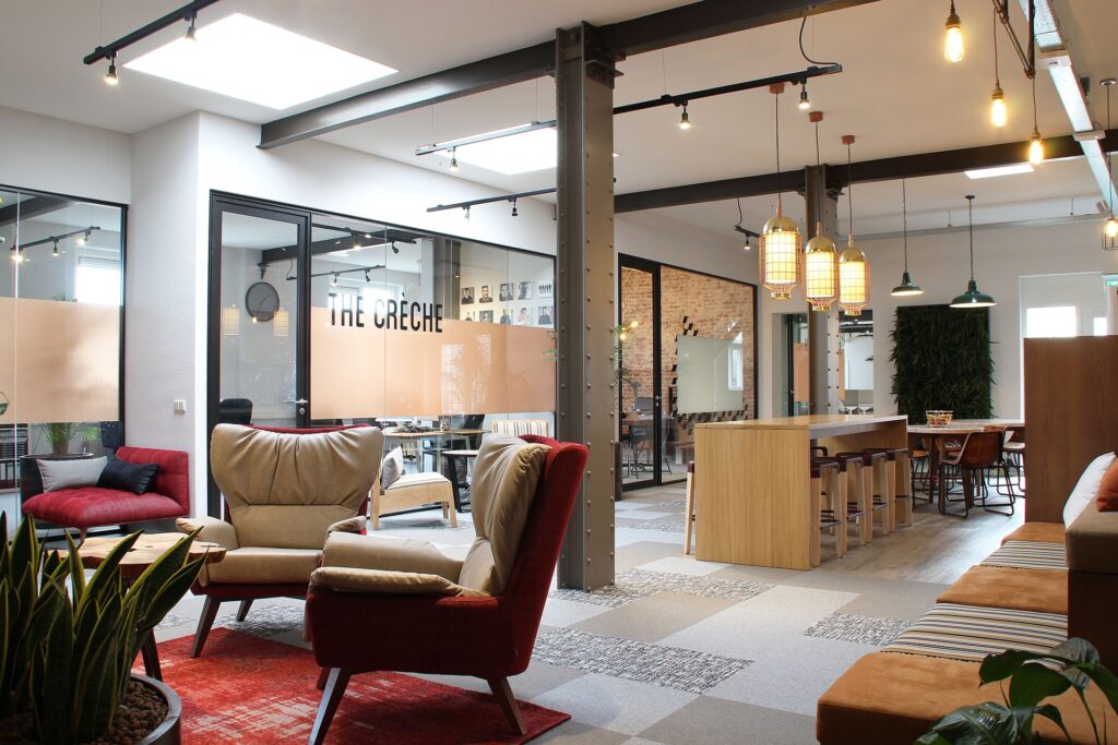 Een moderne kantoorruimte met een gezellige zithoek met fauteuils op de voorgrond. Achter glazen wanden is een vergaderruimte zichtbaar met de tekst “The Crèche” op het glas. Rechts bevindt zich een houten bar met hanglampen en een open werkruimte met ind