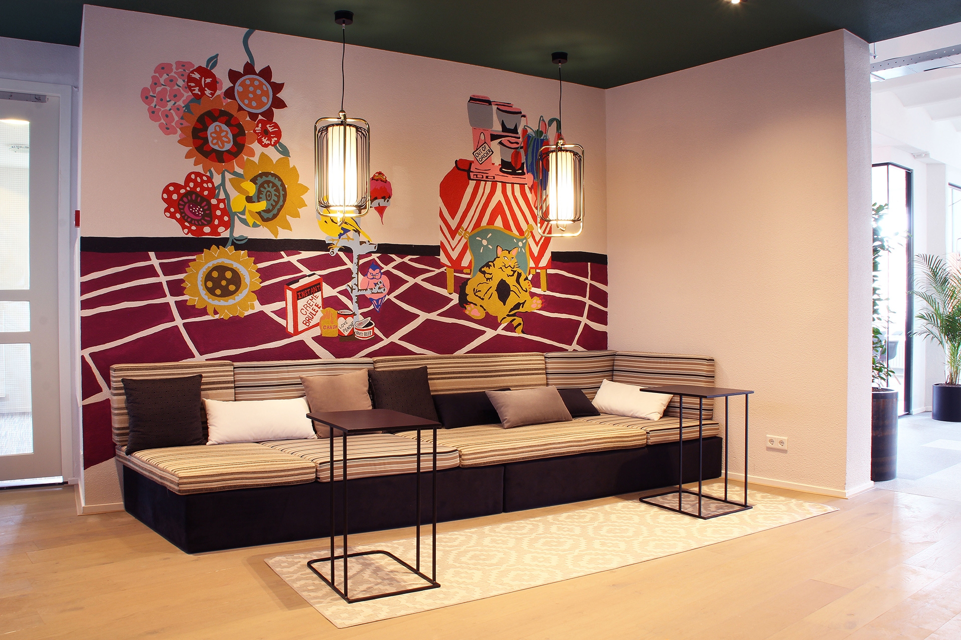 Een moderne zithoek met een lange bank met kussens en twee zwarte bijzettafels op een licht vloerkleed. Aan de muur hangt een kleurrijke muurschildering met tandwielen en illustraties. Boven de bank hangen twee hanglampen en de ruimte heeft een warme, cre