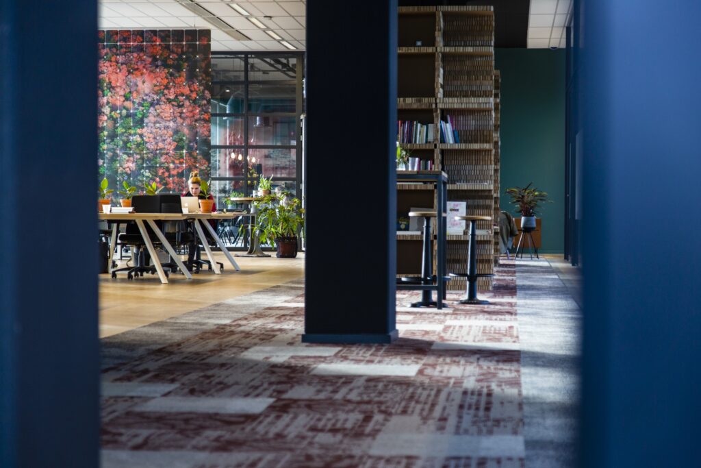 Een modern kantoorinterieur met een open werkruimte, houten tafels en stoelen, planten en een kleurrijke bloemenmuur op de achtergrond. In het midden staat een donkere kolom en rechts is een hoge boekenkast zichtbaar. Het geheel heeft een warme, creatieve