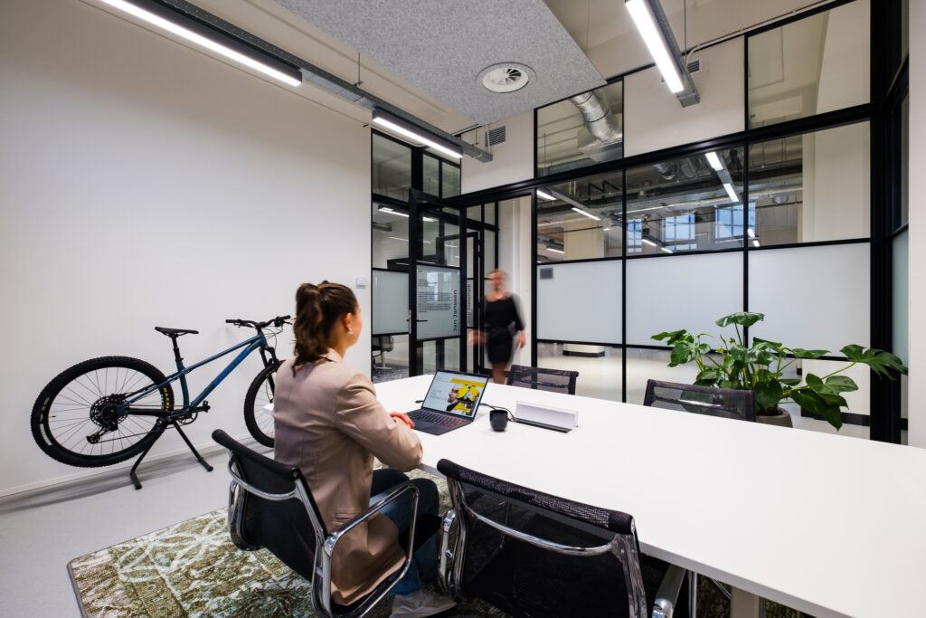 Moderne vergaderruimte met glazen wanden waar een medewerker aan een laptop werkt en een collega binnenloopt.