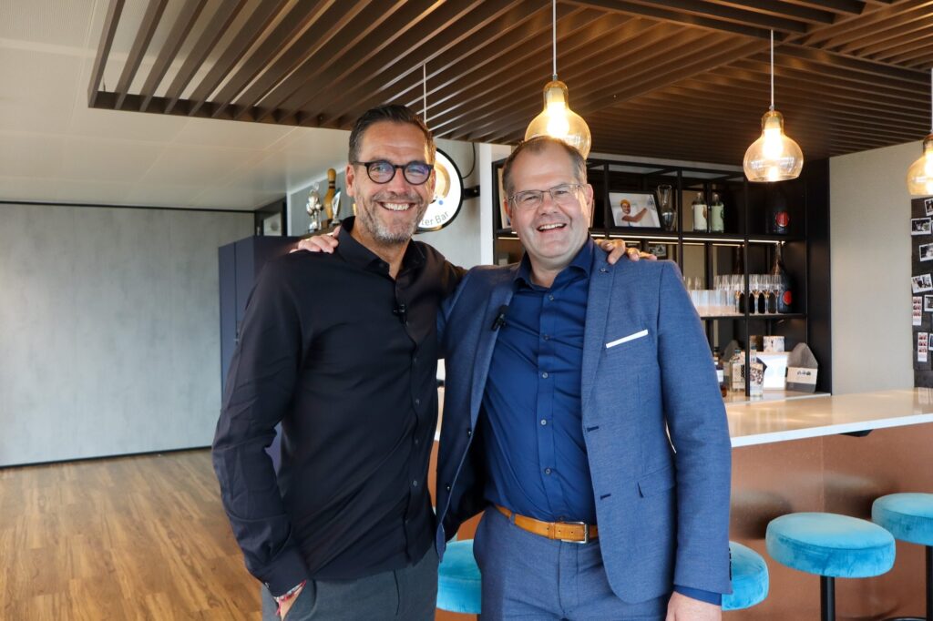 Twee mannen staan lachend naast elkaar in moderne kantooromgeving met bar en hanglampen.
