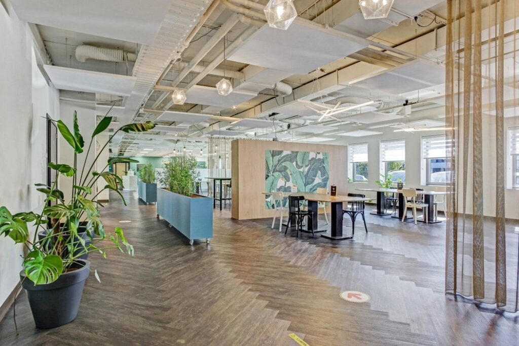 Een ruime, moderne kantoorruimte met een houten visgraatvloer en verschillende werk- en ontmoetingsplekken. In het midden staat een houten wand met een botanische muurschildering. Verspreid door de ruimte staan hoge tafels, plantenbakken en zitplekken. He