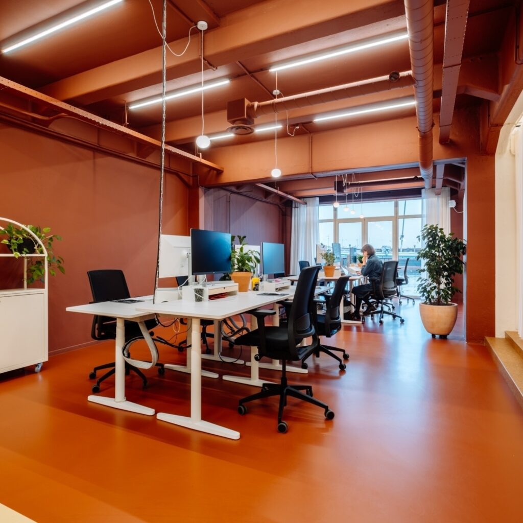 Moderne kantoorruimte met opvallende oranje vloer en plafond, witte bureaus met schermen en groene planten, waar een open en creatieve werkomgeving ontstaat met veel daglicht.