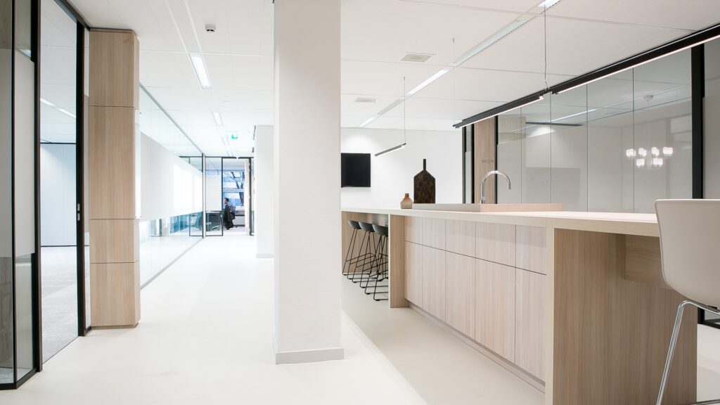 Een lichte, moderne kantoorruimte met een lange houten bar en zwarte barkrukken. Op de bar staat een kraan en enkele decoratieve objecten. Links loopt een gang met glazen vergaderruimtes en aan het einde is een werkplek zichtbaar. De ruimte heeft een stra